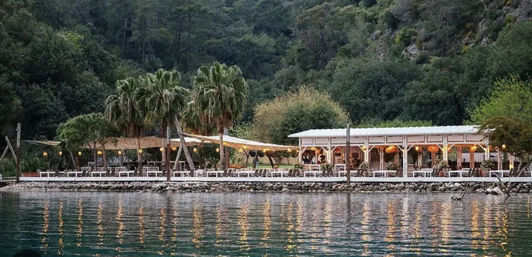 A boat tied to a wooden jetty in an Aegean cove, an evening table set at the water's edge